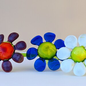 May include: Three glass flower sculptures with green stems. The flowers are in shades of purple, blue, and white. The centre of each flower is a different colour: orange, green, and yellow.