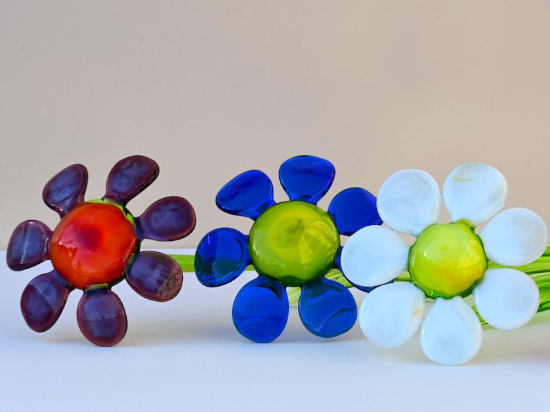 May include: Three glass flower sculptures with green stems. The flowers are in shades of purple, blue, and white. The centre of each flower is a different colour: orange, green, and yellow.