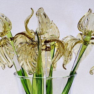 May include: A bouquet of glass irises in a clear glass vase. The flowers are a pale yellow color and have green stems.