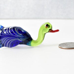 May include: A small glass duck figurine featuring a vibrant blue body with white swirling patterns and a bright lime green neck and head. The duck has a red beak and black eyes, positioned next to a silver US quarter for scale reference.