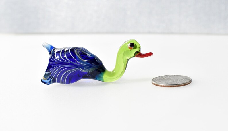 May include: A small glass duck figurine featuring a vibrant blue body with white swirling patterns and a bright lime green neck and head. The duck has a red beak and black eyes, positioned next to a silver US quarter for scale reference.