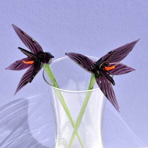 May include: Two glass flower sculptures in a clear vase. The flowers are dark purple with orange centers and green stems. The vase is clear, and the background is a light purple.