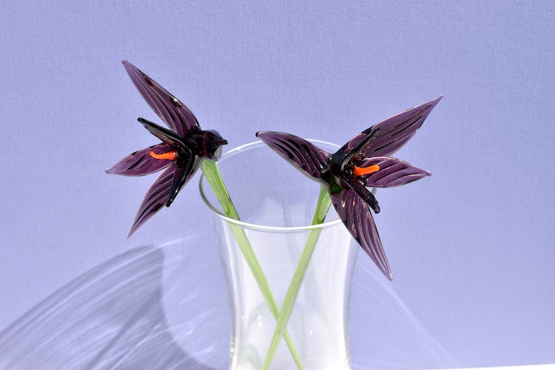 May include: Two glass flower sculptures in a clear vase. The flowers are dark purple with orange centers and green stems. The vase is clear, and the background is a light purple.