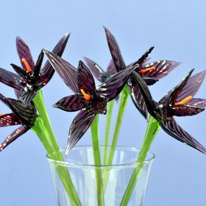 May include: A bouquet of glass flowers in a clear vase. The flowers are a deep purple with orange centers and green stems. The glass petals have a textured appearance, and the vase sits against a light blue background.