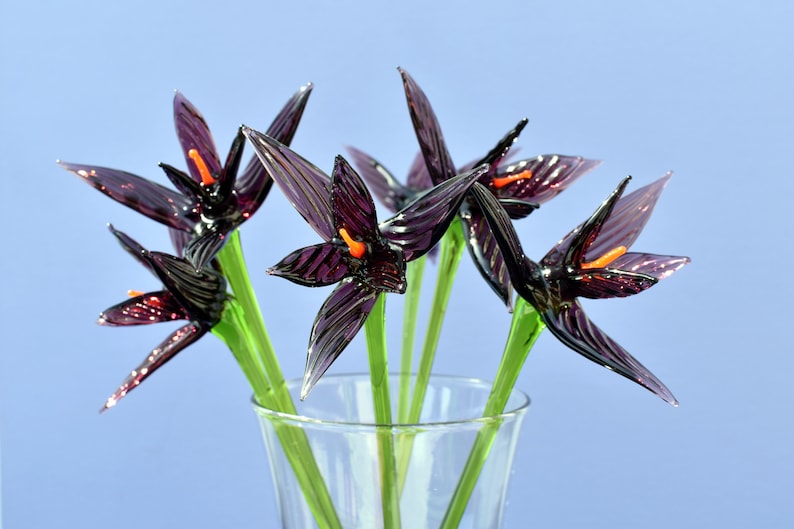 May include: A bouquet of glass flowers in a clear vase. The flowers are a deep purple with orange centers and green stems. The glass petals have a textured appearance, and the vase sits against a light blue background.