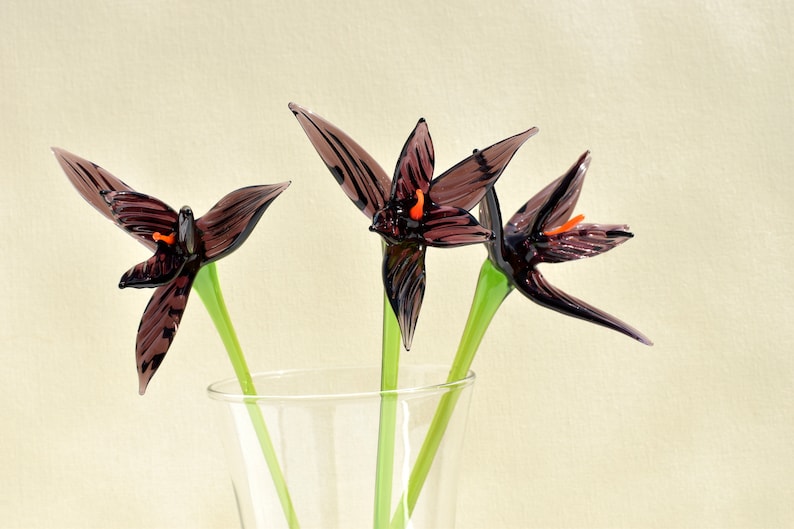 May include: Three glass flowers with dark purple petals and orange accents are displayed in a clear vase. The stems are green. The flowers are in a clear glass vase against a cream-colored background.