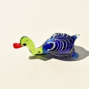 May include: A glass figurine shaped like a duck with a light green neck and head, a bright red beak, and a dark blue body featuring white swirling patterns. The piece has a smooth, glossy finish and is crafted from translucent colored glass.