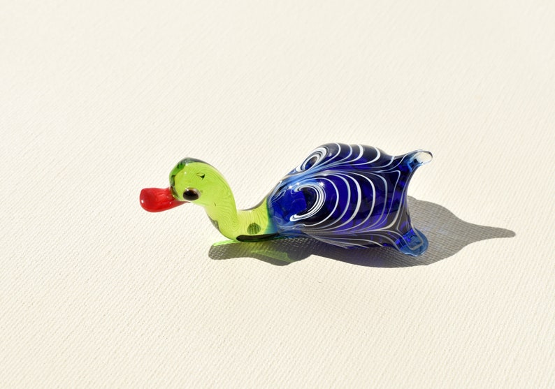 May include: A glass figurine shaped like a duck with a light green neck and head, a bright red beak, and a dark blue body featuring white swirling patterns. The piece has a smooth, glossy finish and is crafted from translucent colored glass.