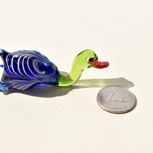 May include: A small glass duck figurine featuring a deep blue body with white swirling patterns, a light green neck, and a red beak. The duck is positioned next to a United States quarter coin for scale, which has a diameter of 0.955 inches.