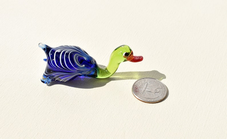 May include: A small glass duck figurine featuring a deep blue body with white swirling patterns, a light green neck, and a red beak. The duck is positioned next to a United States quarter coin for scale, which has a diameter of 0.955 inches.