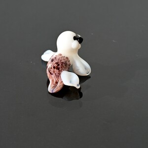 May include: A small, glass seal figurine. The seal has a white body with brown and white markings on its side and flippers. It has black eyes and a black nose. The figurine is on a reflective black surface.
