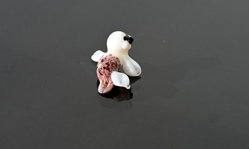 May include: A small, glass seal figurine. The seal has a white body with brown and white markings on its side and flippers. It has black eyes and a black nose. The figurine is on a reflective black surface.