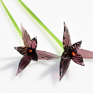May include: Two glass flower sculptures with purple petals and orange centers. The petals have dark purple veining. The stems are green and extend upwards. The flowers are set against a white background.