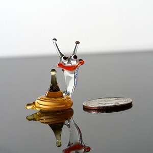 May include: A small glass snail figurine with a yellow shell and a clear glass body. The snail has red eyes and a red mouth. The figurine is sitting on a reflective surface.