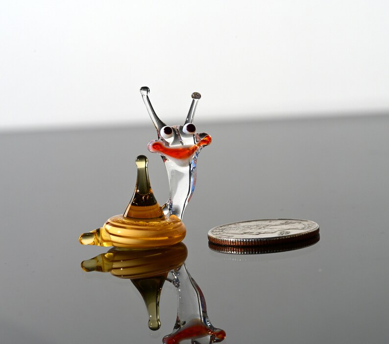 May include: A small glass snail figurine with a yellow shell and a clear glass body. The snail has red eyes and a red mouth. The figurine is sitting on a reflective surface.