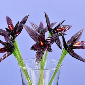 May include: A glass vase holding a bouquet of handcrafted glass flowers. The flowers are a deep purple with orange centers and green stems. The petals are pointed and have a textured appearance. The vase is clear, and the background is a solid light blue.