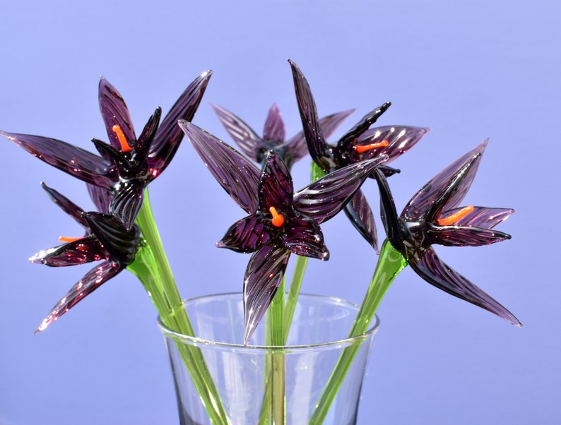 May include: A glass vase holding a bouquet of handcrafted glass flowers. The flowers are a deep purple with orange centers and green stems. The petals are pointed and have a textured appearance. The vase is clear, and the background is a solid light blue.