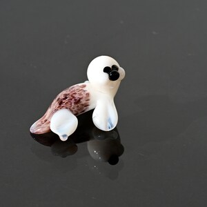 May include: A small, glass seal figurine. The seal has a white body with brown and purple markings on its back and flippers. It has black eyes and a black nose. The figurine is sitting on a reflective surface.