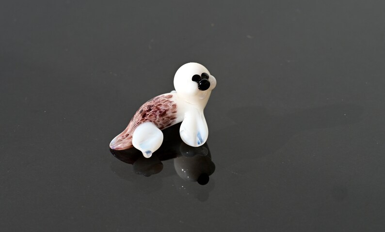 May include: A small, glass seal figurine. The seal has a white body with brown and purple markings on its back and flippers. It has black eyes and a black nose. The figurine is sitting on a reflective surface.