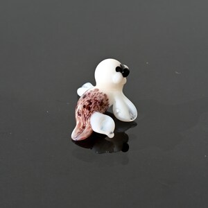 May include: A small, handcrafted glass seal figurine. The seal has a white body, black eyes, and a brown and white patterned tail. The figurine is posed in a sitting position, with its flippers extended. The glass is translucent, allowing light to pass through.
