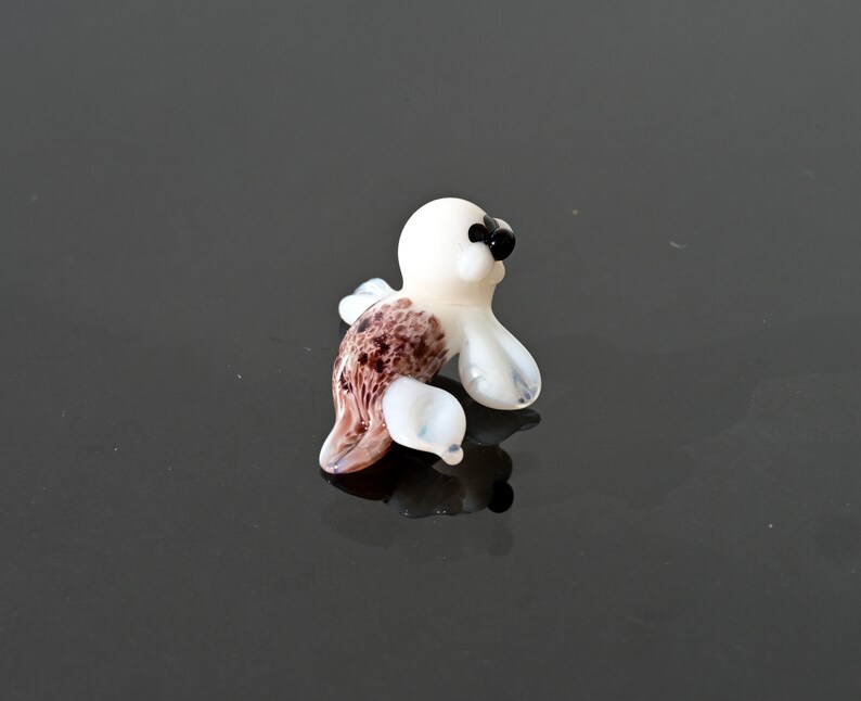 May include: A small, handcrafted glass seal figurine. The seal has a white body, black eyes, and a brown and white patterned tail. The figurine is posed in a sitting position, with its flippers extended. The glass is translucent, allowing light to pass through.