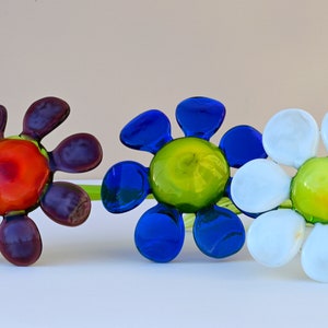 May include: Three glass flower sculptures with green stems. The flowers are in shades of purple, blue, and white. The centre of each flower is a different colour: red, green, and green.