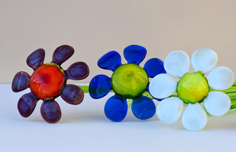 May include: Three glass flower sculptures with green stems. The flowers are in shades of purple, blue, and white. The centre of each flower is a different colour: red, green, and green.