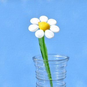 May include: A glass daisy flower with a yellow center and white petals in a clear glass vase with a textured surface.
