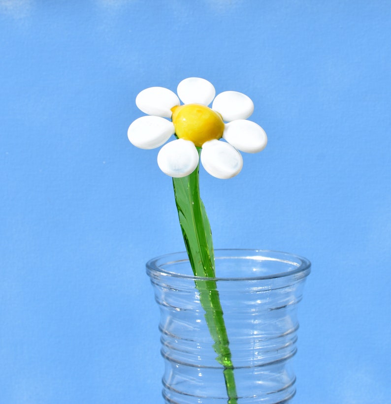 May include: A glass daisy flower with a yellow center and white petals in a clear glass vase with a textured surface.