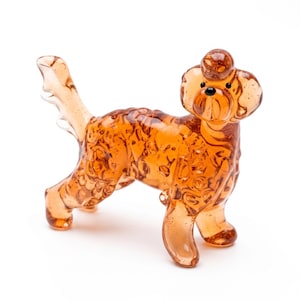 Puede incluir: Una figura de cristal de color ámbar translúcido que representa a un perro de pie de perfil. La escultura presenta una superficie texturizada con burbujas, una cola levantada y un copete distintivo en la cabeza. El perro tiene ojos y nariz de cristal redondos y oscuros.