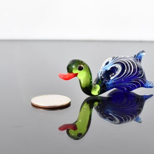 May include: A small glass duck figurine featuring a green head, a bright red beak, and a dark blue body with intricate white swirling patterns. The duck is positioned next to a silver-colored coin for scale, showing its miniature size.