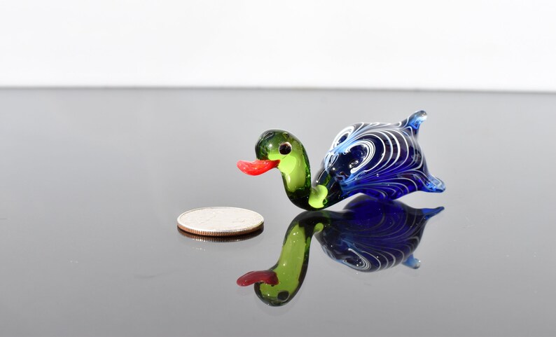 May include: A small glass duck figurine featuring a green head, a bright red beak, and a dark blue body with intricate white swirling patterns. The duck is positioned next to a silver-colored coin for scale, showing its miniature size.