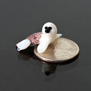 May include: A miniature glass seal figurine, primarily white with a speckled brown tail, rests on a quarter. The seal has black eyes and a black nose. The figurine is a handcrafted item.