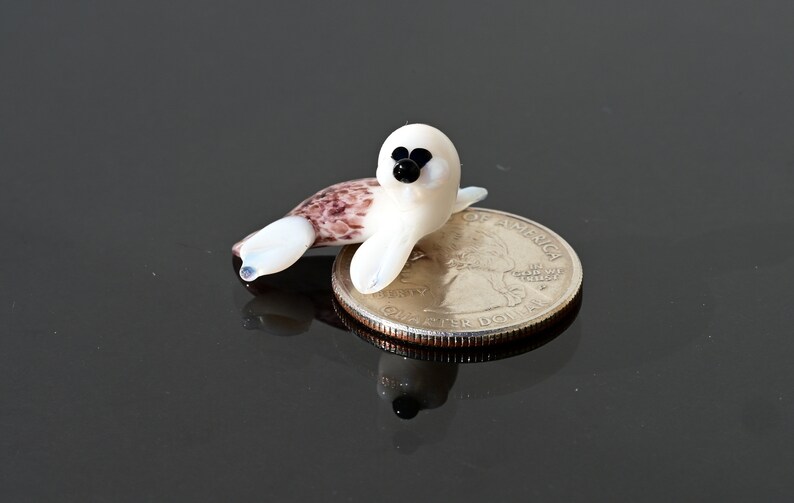 May include: A miniature glass seal figurine, primarily white with a speckled brown tail, rests on a quarter. The seal has black eyes and a black nose. The figurine is a handcrafted item.