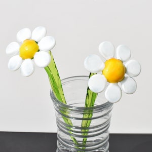May include: Two white glass daisies with yellow centers in a clear glass vase with a textured pattern. The stems are green glass.