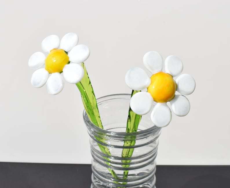 May include: Two white glass daisies with yellow centers in a clear glass vase with a textured pattern. The stems are green glass.