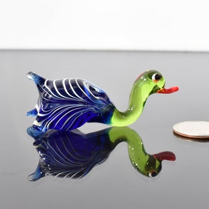 May include: A small glass duck figurine featuring a deep blue body with white swirling patterns, a light green neck and head, and a bright orange beak. A standard US quarter coin is placed next to the figurine to provide a sense of scale.