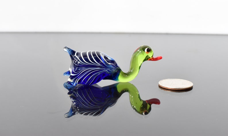 May include: A small glass duck figurine featuring a deep blue body with white swirling patterns, a light green neck and head, and a bright orange beak. A standard US quarter coin is placed next to the figurine to provide a sense of scale.