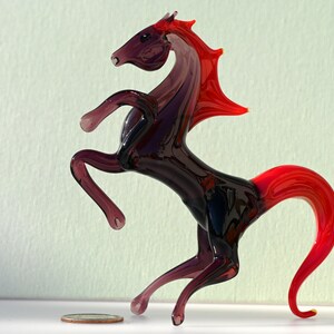 Peut inclure: Une figurine de cheval en verre avec une crini&egrave;re et une queue rouges. Le cheval est debout sur ses pattes arri&egrave;re, ses pattes avant lev&eacute;es. La figurine est en verre transparent avec une teinte violette.