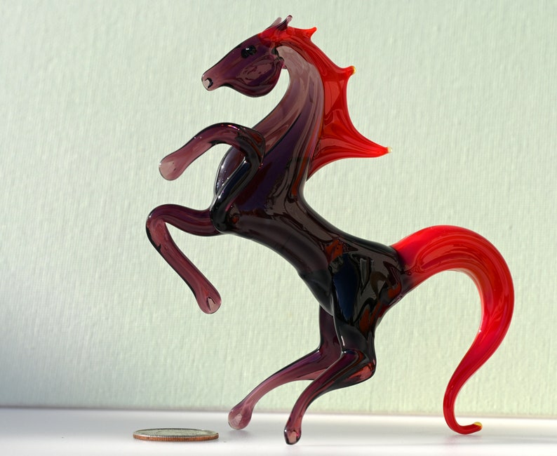 Peut inclure: Une figurine de cheval en verre avec une crini&egrave;re et une queue rouges. Le cheval est debout sur ses pattes arri&egrave;re, ses pattes avant lev&eacute;es. La figurine est en verre transparent avec une teinte violette.