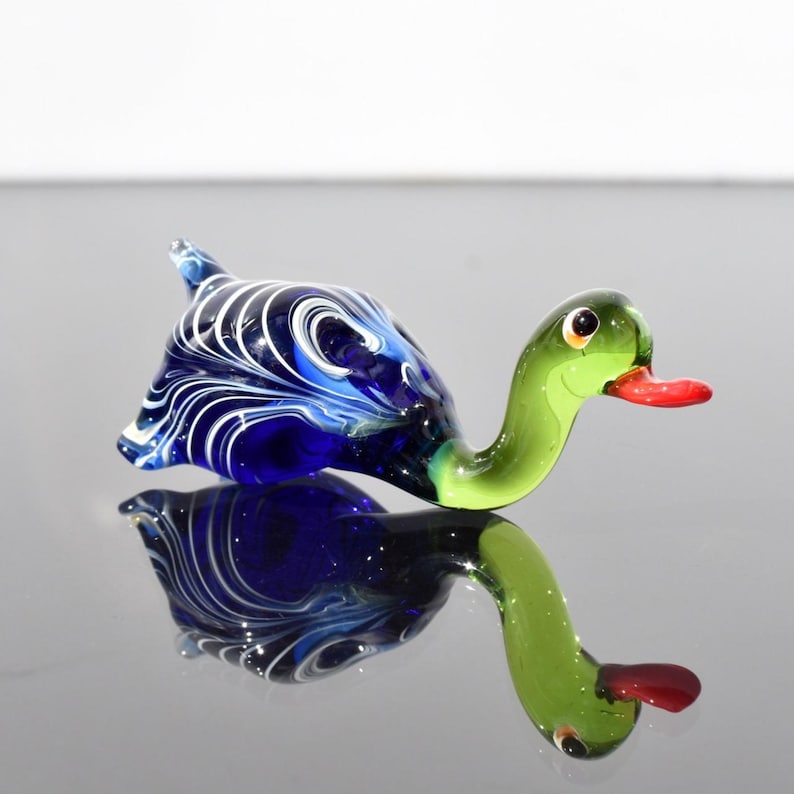 May include: A glass figurine shaped like a duck with a deep blue body featuring white swirling patterns. The neck and head are a translucent light green, and the beak is a bright red. The figurine has a smooth, glossy finish throughout.