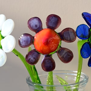 May include: Three glass flowers in a clear vase. The flowers are white, purple, and blue with green stems. The centre of the purple flower is orange.