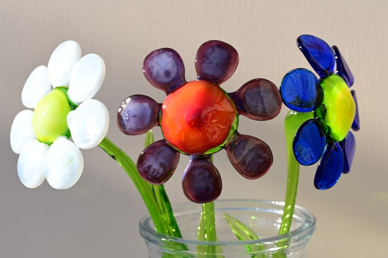 May include: Three glass flowers in a clear vase. The flowers are white, purple, and blue with green stems. The centre of the purple flower is orange.