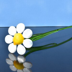 May include: A glass daisy flower with a yellow center and white petals on a green stem. The flower is laying on a blue surface.