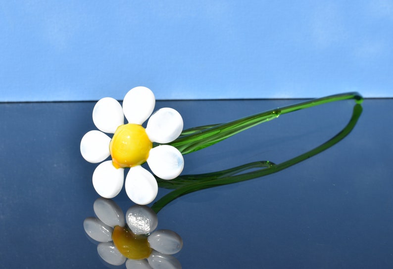 May include: A glass daisy flower with a yellow center and white petals on a green stem. The flower is laying on a blue surface.