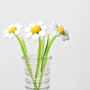 May include: Three white glass daisies with yellow centers in a clear glass vase. The vase has a textured design.