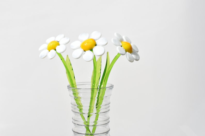 May include: Three white glass daisies with yellow centers in a clear glass vase. The vase has a textured design.
