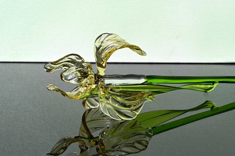 May include: A glass flower sculpture with a yellow flower and green stem. The flower is made of glass and has a delicate, intricate design. The stem is also made of glass and is a vibrant green color.