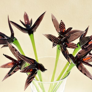May include: A bouquet of glass flowers in a clear vase. The flowers are dark purple with orange accents and green stems. The petals are pointed and have a striped pattern. The vase is partially visible, and the background is a light beige.