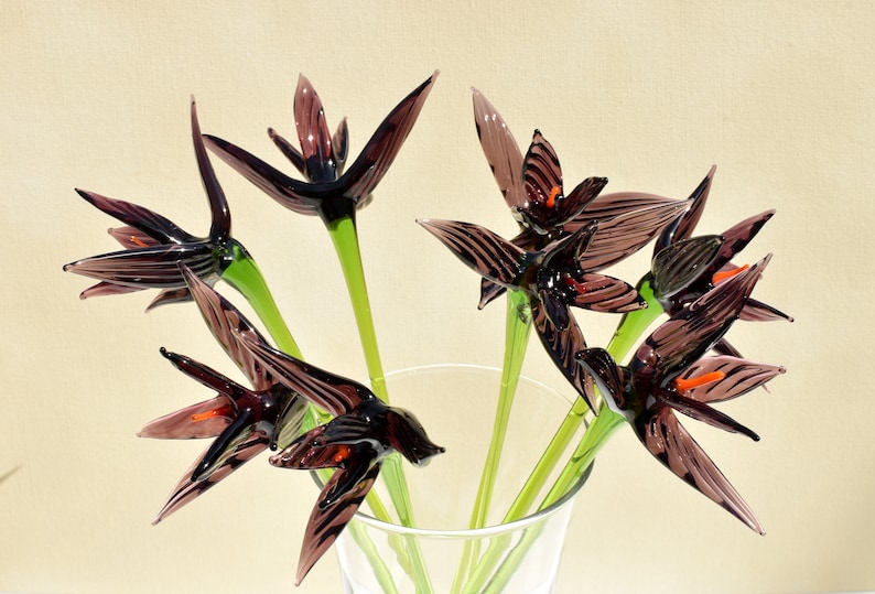 May include: A bouquet of glass flowers in a clear vase. The flowers are dark purple with orange accents and green stems. The petals are pointed and have a striped pattern. The vase is partially visible, and the background is a light beige.